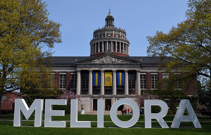 Large white letters spelling MELIORA standing on the Eastman Quad in from of Rush Rhees Library.