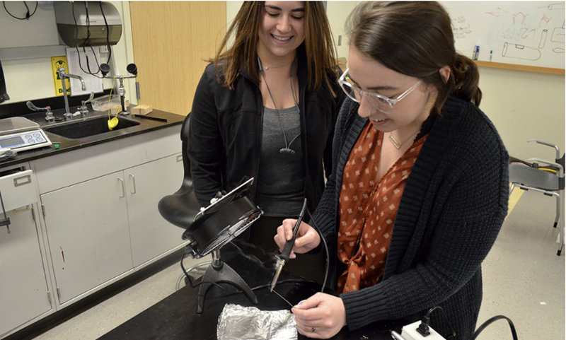 From left, Rebekah Abrams ’19 and Taryn Milnes ’19 demonstrate a prototype of a fume absorber they designed for a Unistel assembly line that employs workers with developmental disabilities. (University of Rochester photo / Bob Marcotte)