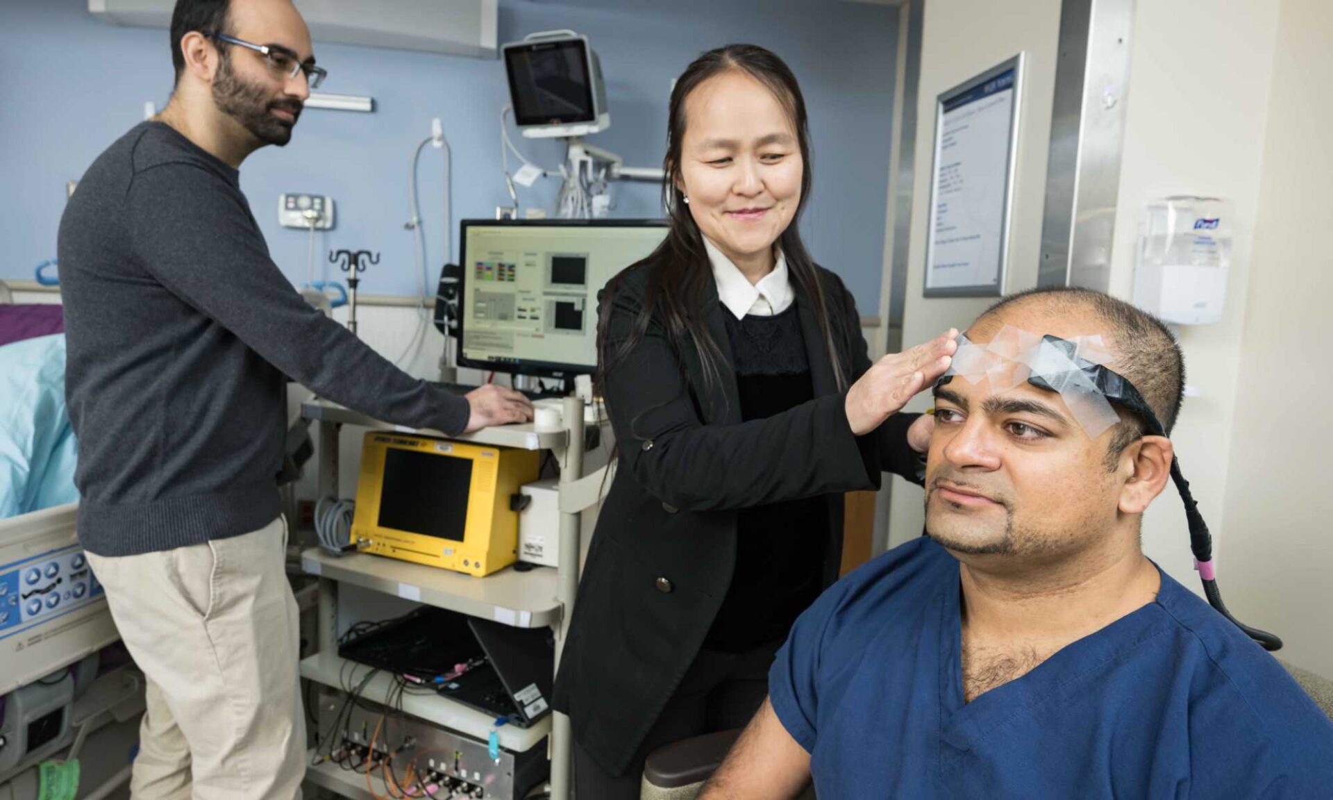 Two researchers and a participant demonstrate how multiple devices linked together can evaluate different aspects of the brain’s health during extracorporeal membrane oxygenation (ECMO) therapy.