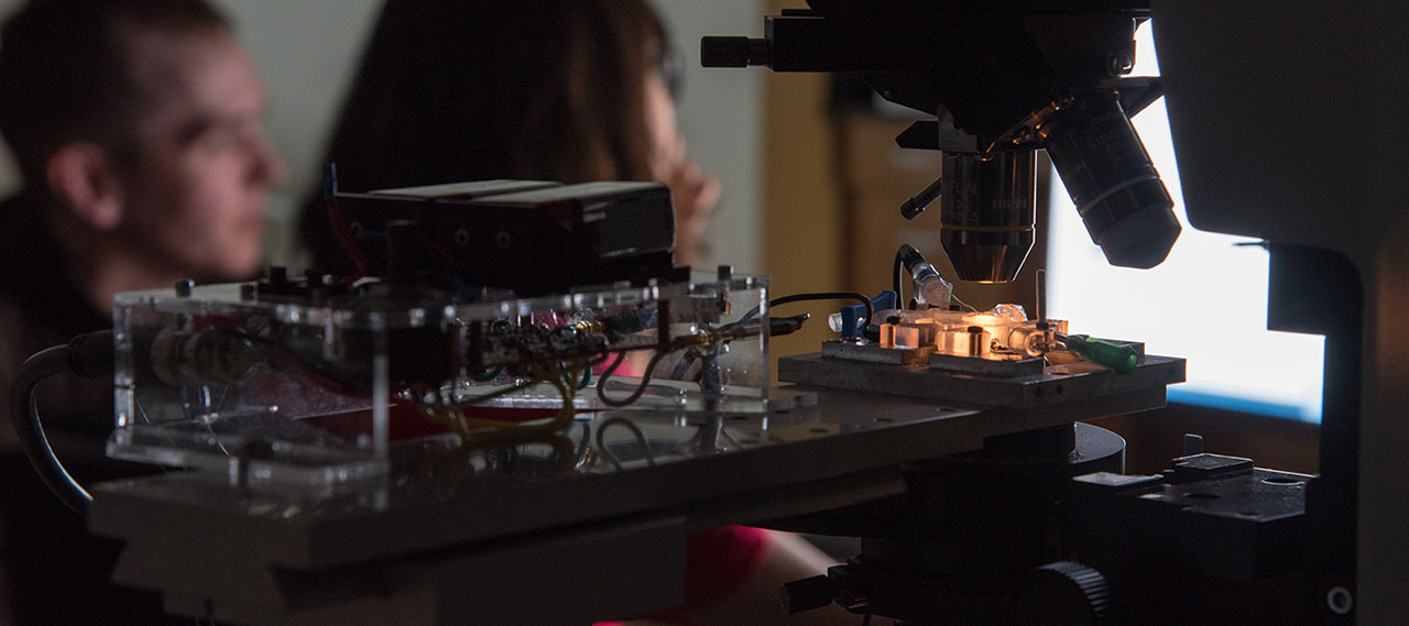 A close up of a microscope with 2 students in the background.