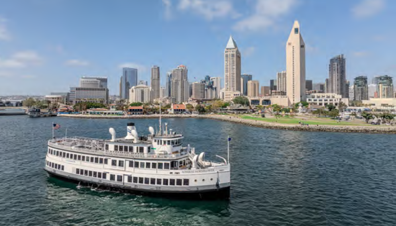A small cruise ship on water near a shore with a cityscape in the background.