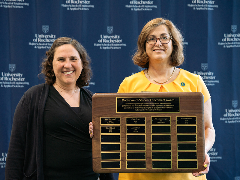 Two people smile at the camera while the one on the right holds a plaque.