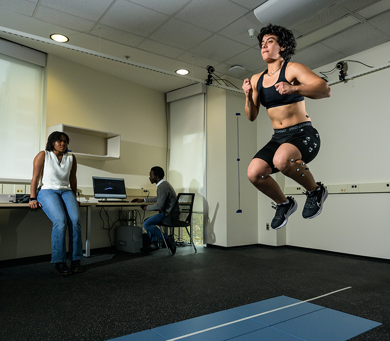 A student performs various movement and balance tasks on a force plate walkway while wearing shoes instrumented with pressure insoles and a suit outfitted with reflective markers for use in collection of biomechanics data as two other students look on.