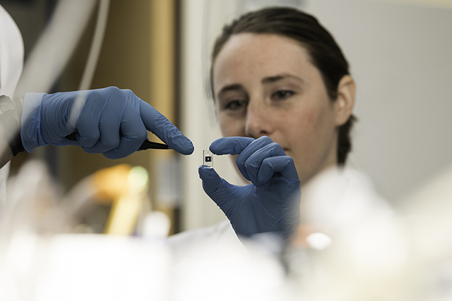 A gloved hand holding a microchip while another gloved hand comes at it with a tool. A person's face in the background.