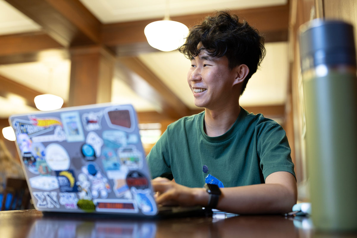 Student in green t shirt works on laptop with stickers on it.