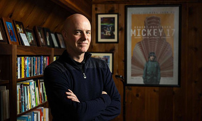 man staring at camera in a room with bookshelves behind him.