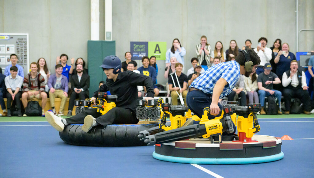 Two seniors demonstrate their project: a disc-like floating vehicle, to an audience of excited colleagues.